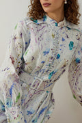Vrouw draagt een lange witte maxi-jurk met een blauwe en lila bloemenprint, lange mouwen brede manchet met 6 knopen en een bijpassende ceintuur. 