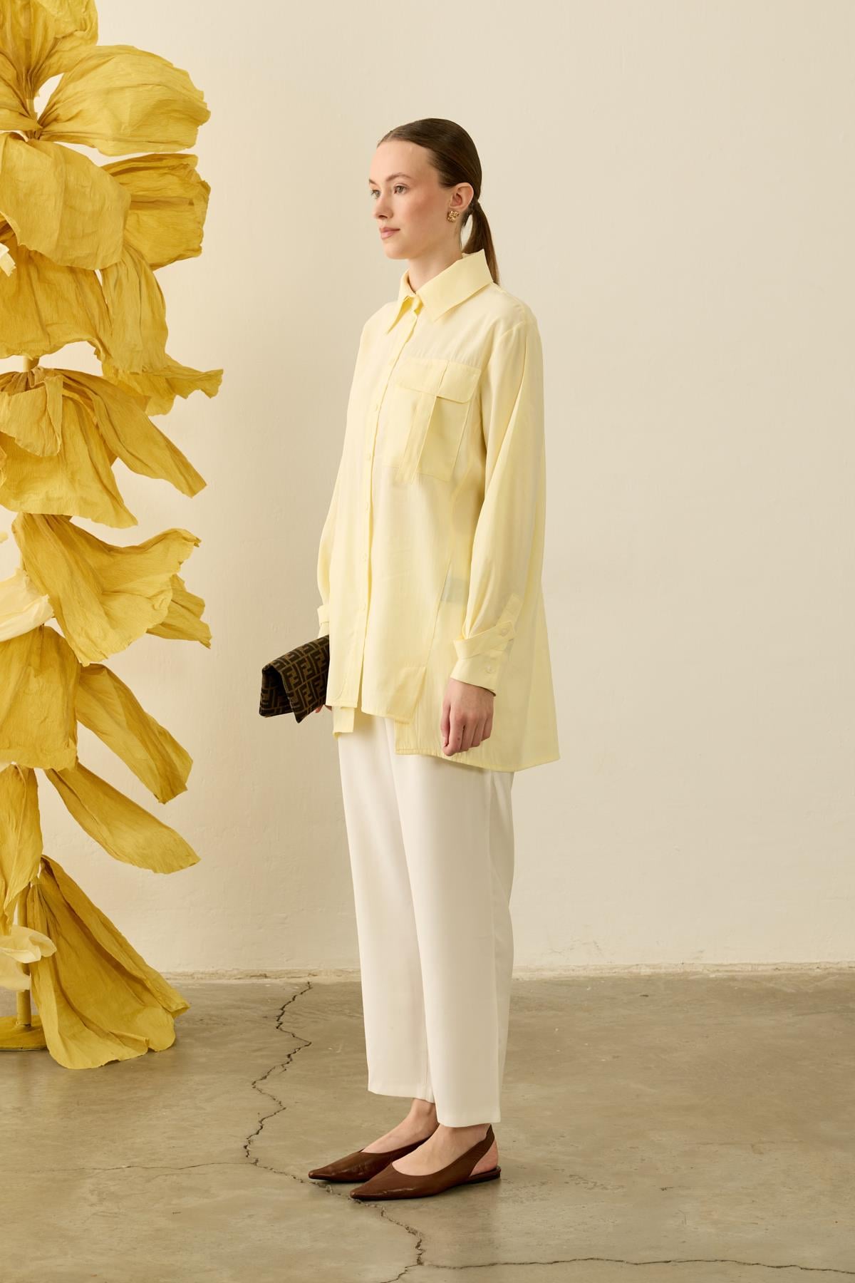 Een zijdezachte, zachtgele (Soft Lemon) tuniekblouse met een soepele valling. De blouse heeft een klassieke puntkraag, een minimalistische blinde knoopsluiting en een opvallende borstzak met utility-detail aan de rechterkant. Het verlengde model is gecombineerd met een witte pantalon en bruine puntige slingback-schoenen.