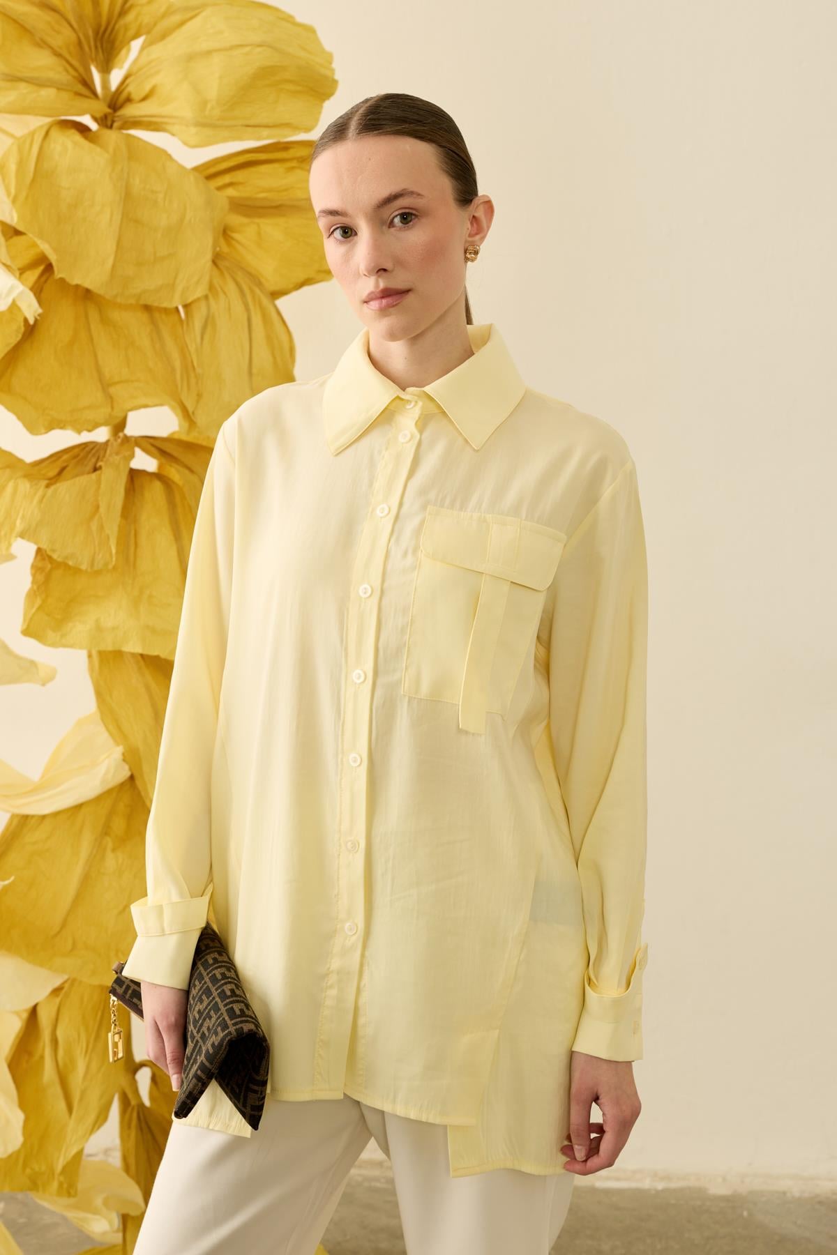 Een zijdezachte, zachtgele (Soft Lemon) tuniekblouse met een soepele valling. De blouse heeft een klassieke puntkraag, een minimalistische blinde knoopsluiting en een opvallende borstzak met utility-detail aan de rechterkant. Het verlengde model is gecombineerd met een witte pantalon.