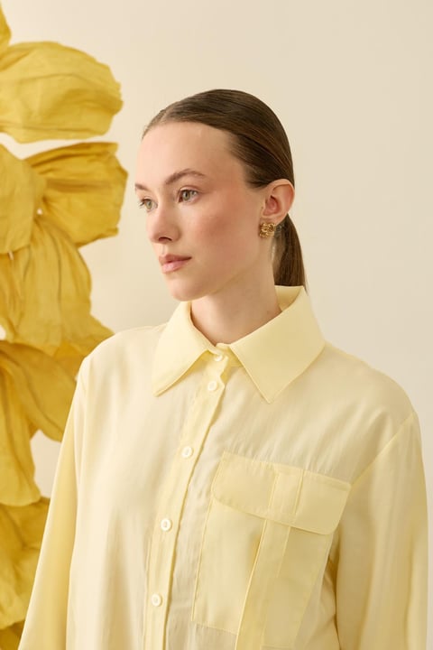 Een zijdezachte, zachtgele (Soft Lemon) tuniekblouse met een soepele valling. De blouse heeft een klassieke puntkraag, een minimalistische blinde knoopsluiting en een opvallende borstzak met utility-detail aan de rechterkant. 