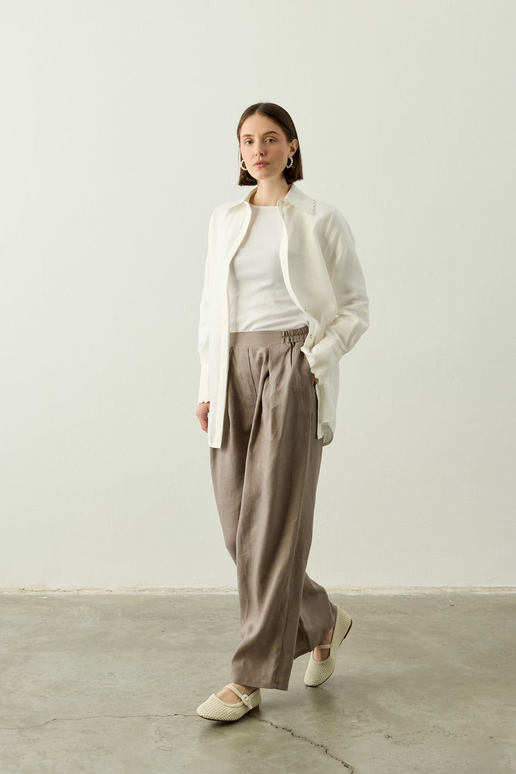 Een model met een bobkapsel poseert in een minimalistische setting met een witte achtergrond en betonnen vloer. Ze draagt de Stone Taupe linnen palazzo pantalon van Atelier SHÉ & CO., gecombineerd met een witte top en een opengevallen witte linnen blouse. Ze draagt ecru gevlochten Mary Jane schoenen en subtiele zilveren hoepeloorbellen. Haar handen zitten nonchalant in de zakken van de broek.