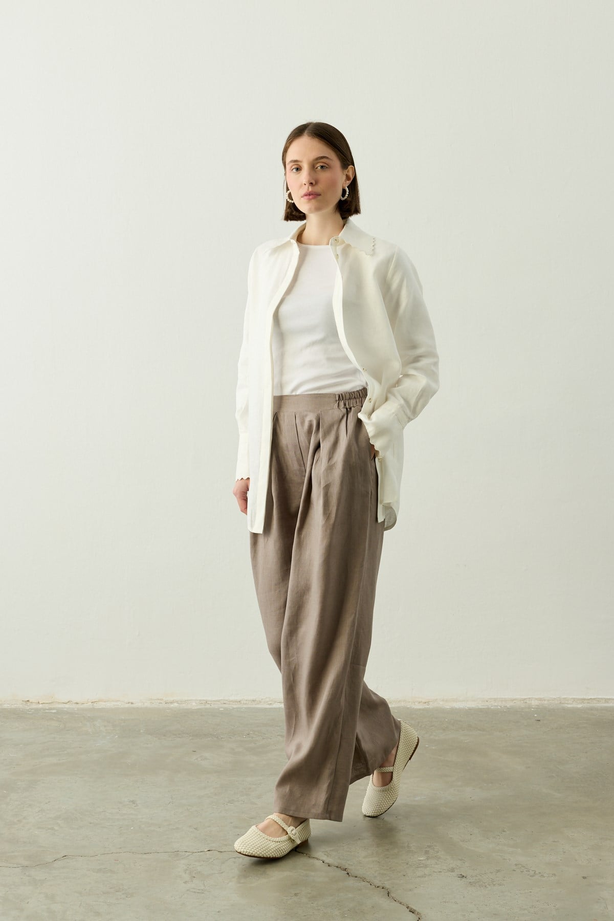 Een model met een bobkapsel poseert in een minimalistische setting met een witte achtergrond en betonnen vloer. Ze draagt de Stone Taupe linnen palazzo pantalon van Atelier SHÉ & CO., gecombineerd met een witte top en een opengevallen witte linnen blouse. Ze draagt ecru gevlochten Mary Jane schoenen en subtiele zilveren hoepeloorbellen. Haar handen zitten nonchalant in de zakken van de broek.
