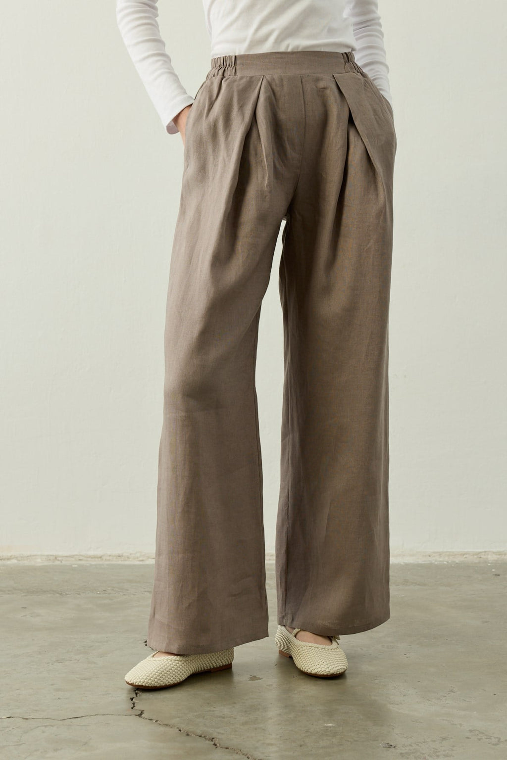 Een close-up van de Mara Trousers in Stone Taupe van Atelier SHÉ & CO. De afbeelding toont een 100% linnen palazzo pantalon met een hoge taille, diepe dubbele plooien aan de voorzijde en een comfortabele elastische tailleband aan de zijkant. De broek heeft wijde pijpen en wordt gedragen met een witte top en ecru gevlochten Mary Jane schoenen op een betonnen vloer.