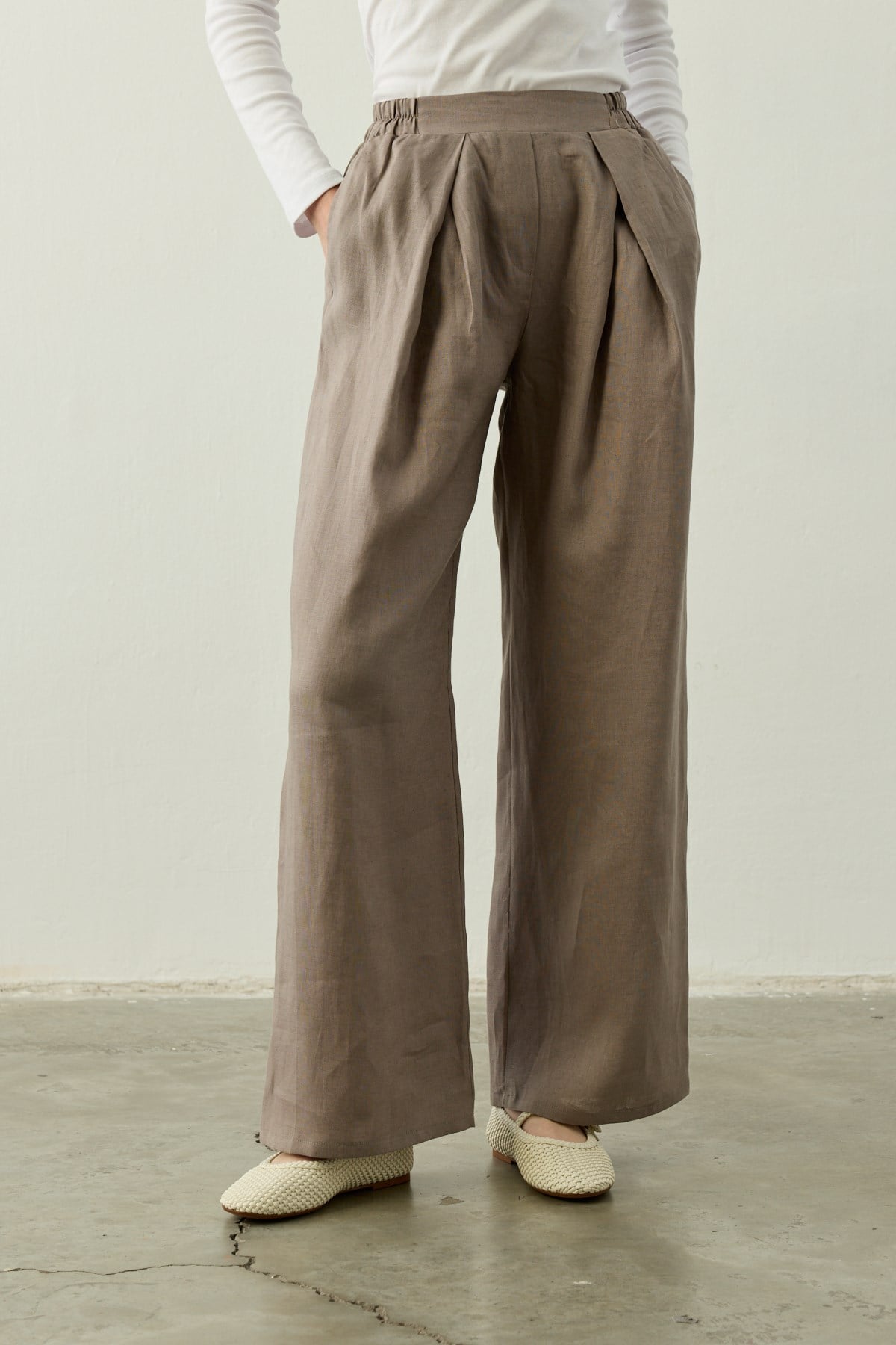 Een close-up van de Mara Trousers in Stone Taupe van Atelier SHÉ & CO. De afbeelding toont een 100% linnen palazzo pantalon met een hoge taille, diepe dubbele plooien aan de voorzijde en een comfortabele elastische tailleband aan de zijkant. De broek heeft wijde pijpen en wordt gedragen met een witte top en ecru gevlochten Mary Jane schoenen op een betonnen vloer.