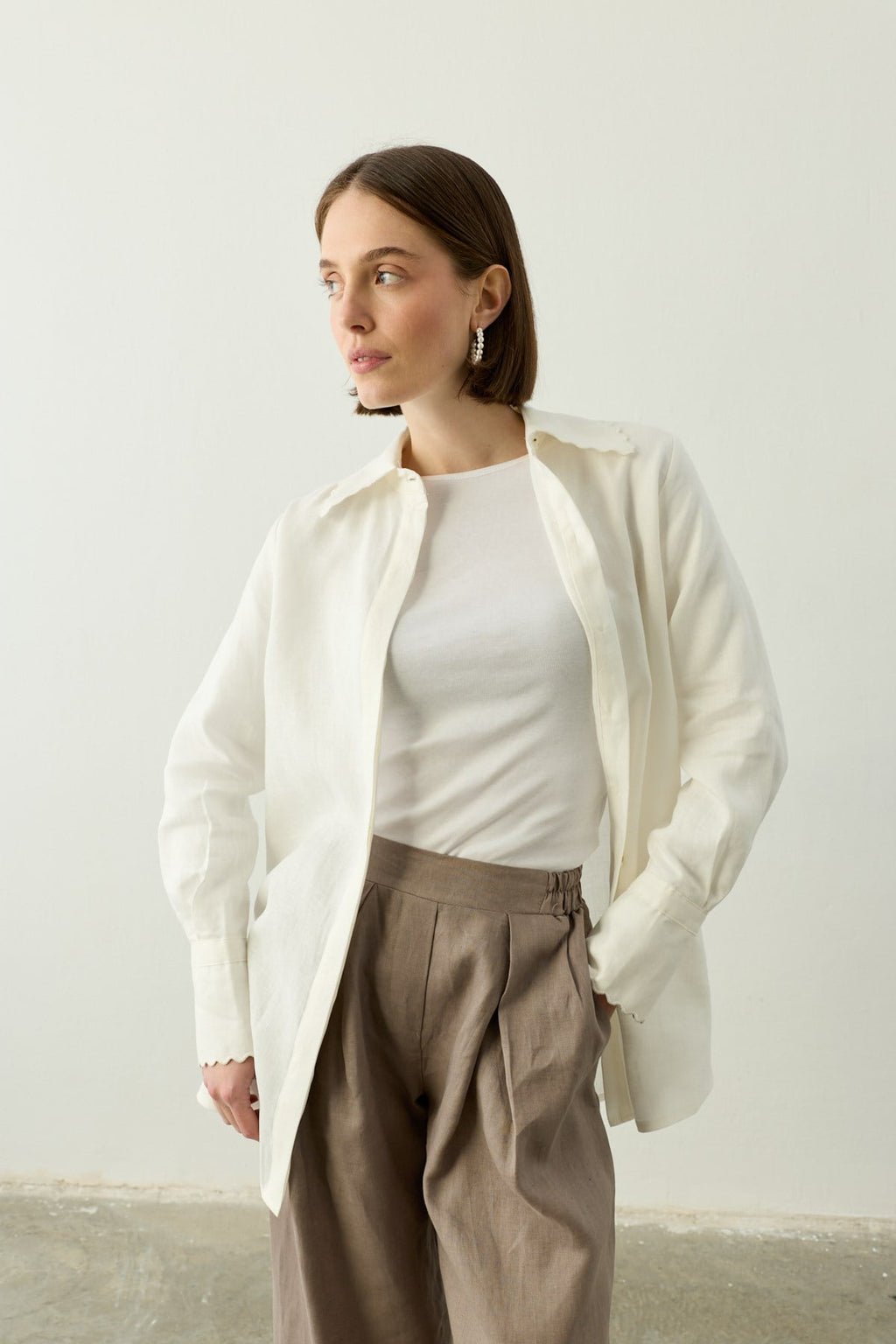 Een close-up van een model dat de linnen "Mara Trousers" in Stone Taupe draagt van Atelier SHÉ & CO. De focus ligt op de details van de broek, zoals de diepe dubbele plooien aan de voorzijde en de gladde tailleband met elastische inzet. Ze draagt de pantalon met een witte top en een losvallende witte linnen blouse met geschulpte randen aan de kraag en manchetten. Het model heeft een kort bruin kapsel en draagt parelmoer-achtige hoepeloorbellen.