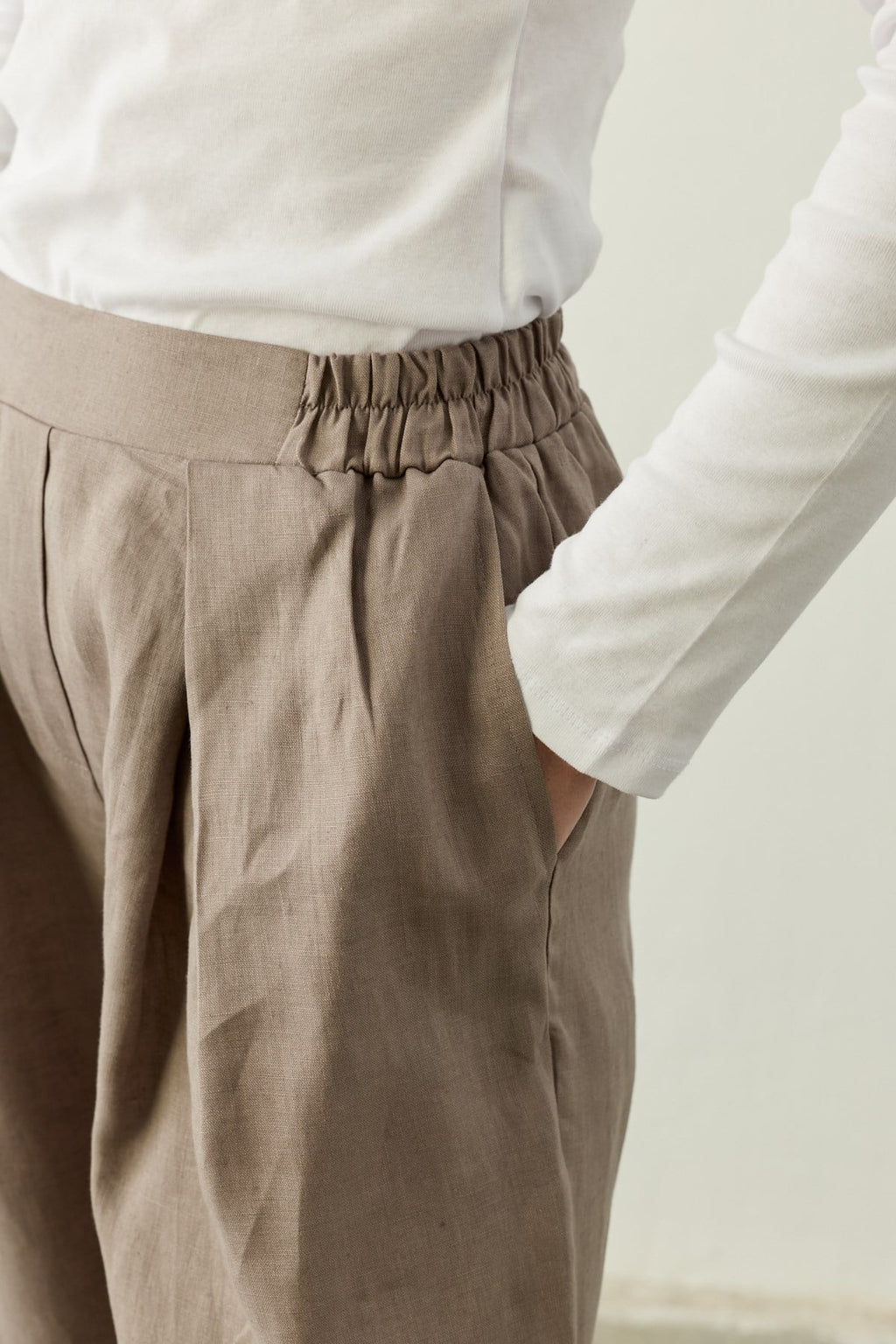 Een gedetailleerde close-up van de tailleband van de "Mara Trousers" in Stone Taupe. De afbeelding brengt de rijke textuur van het 100% pure linnen prachtig in beeld. Te zien is de overgang van de gladde voorzijde van de tailleband naar de elastische inzet aan de zijkant, de diepe dubbele plooien (pleats) en de discreet weggewerkte zijzak waar een hand in rust. De pantalon wordt gedragen met een nauwsluitende witte top.