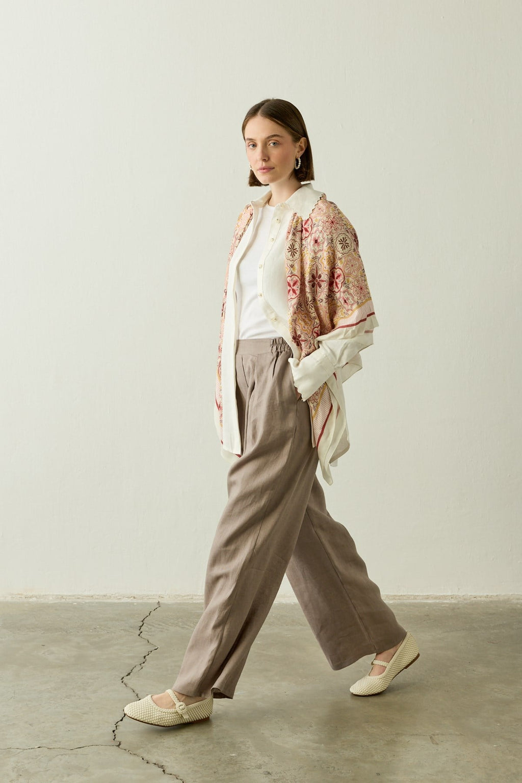 Een zij-aanzicht van een model dat loopt, gekleed in de "Mara Trousers" in Stone Taupe van Atelier SHÉ & CO. De ruimvallende palazzo pantalon van 100% puur linnen is gecombineerd met een witte top en een opengevallen crèmekleurige blouse. Over de schouders hangt een sjaal met een gedetailleerd bloemen- en tegelmotief in roze en geel. Het model draagt ecru gevlochten instappers.
