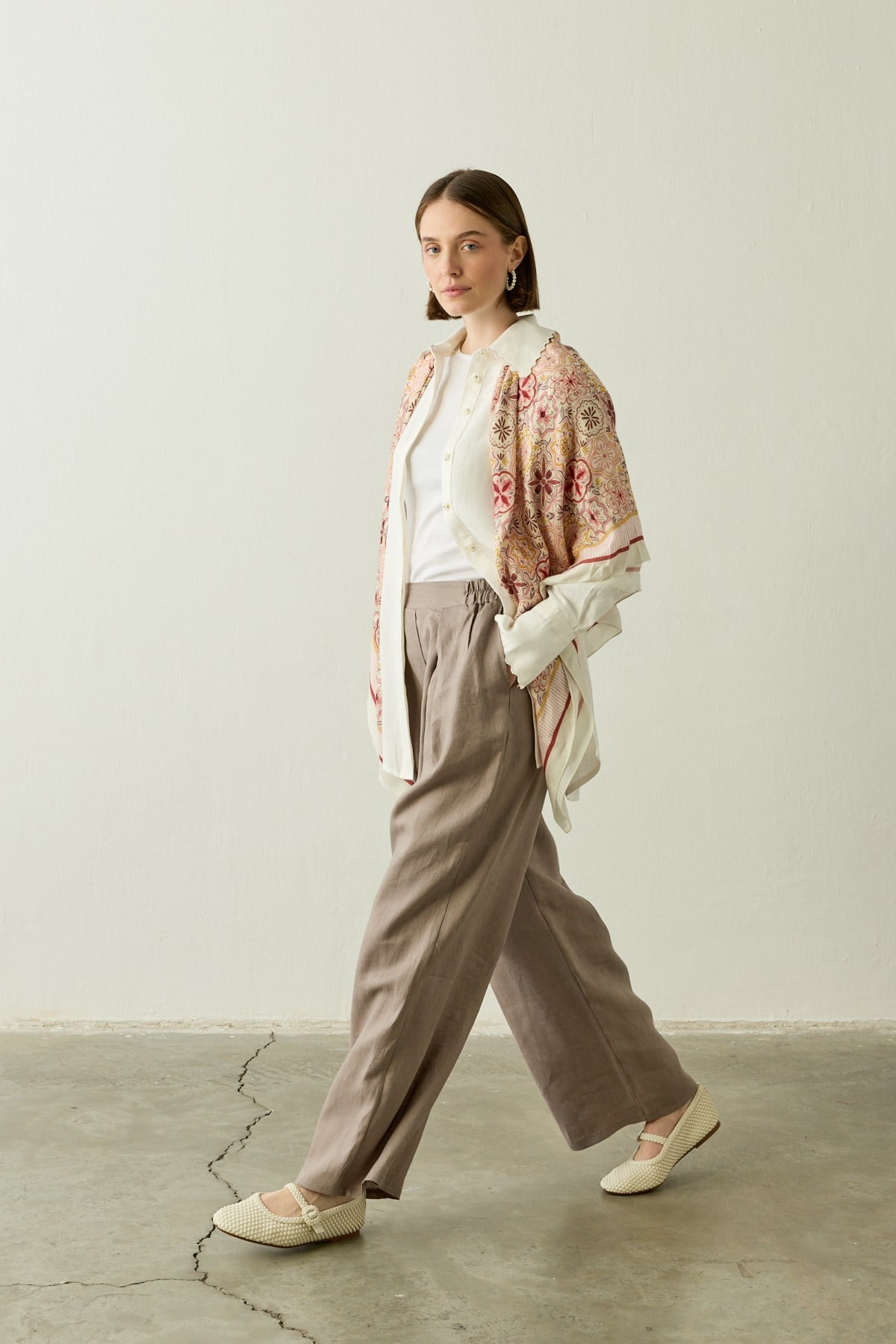 Een zij-aanzicht van een model dat loopt, gekleed in de "Mara Trousers" in Stone Taupe van Atelier SHÉ & CO. De ruimvallende palazzo pantalon van 100% puur linnen is gecombineerd met een witte top en een opengevallen crèmekleurige blouse. Over de schouders hangt een sjaal met een gedetailleerd bloemen- en tegelmotief in roze en geel. Het model draagt ecru gevlochten instappers.