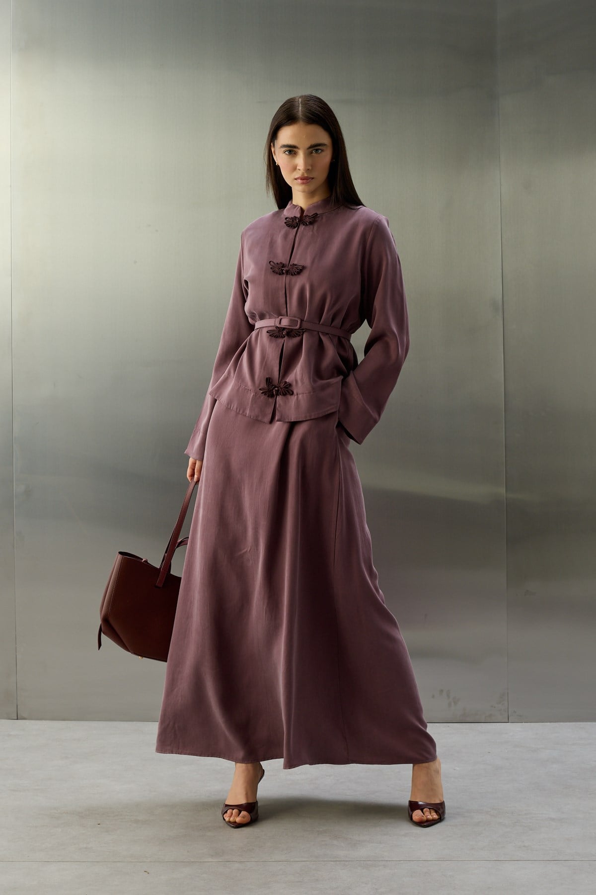 Een volledige lichaamsfoto van een model dat een paarse, zijdeachtige tweedelige set draagt, bestaande uit een getailleerd jasje met een riem en een lange rok. Het jasje heeft een opstaande kraag en is aan de voorzijde versierd met drie donkerpaarse, sierlijke vlindersluitingen. De rok valt soepel tot op de enkels. Het model houdt een bordeauxrode tas vast en draagt bijpassende open hakken. De achtergrond is van industrieel geborsteld metaal.