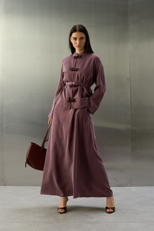 Een volledige lichaamsfoto van een model dat een paarse, zijdeachtige tweedelige set draagt, bestaande uit een getailleerd jasje met een riem en een lange rok. Het jasje heeft een opstaande kraag en is aan de voorzijde versierd met drie donkerpaarse, sierlijke vlindersluitingen. De rok valt soepel tot op de enkels. Het model houdt een bordeauxrode tas vast en draagt bijpassende open hakken. De achtergrond is van industrieel geborsteld metaal.