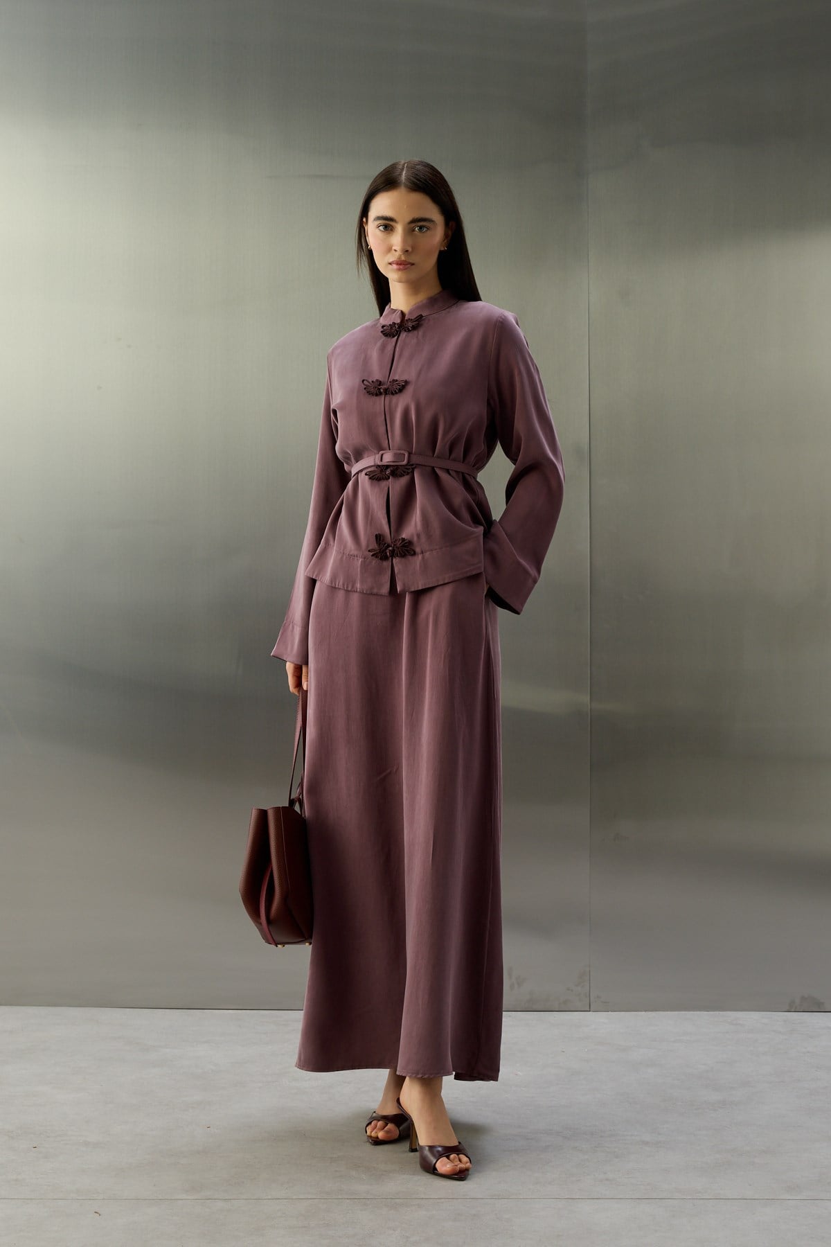 Een full-body opname van een model tegen een minimalistische, grijs-metalen achtergrond. Ze draagt een elegante, paarse tweedelige set van een gladde, zijdeachtige stof. Het jasje heeft een opstaande kraag, lange mouwen en is voorzien van drie donkere, sierlijke knoopsluitingen (kikersluitingen) aan de voorzijde. Een bijpassende riem met gesp accentueert de taille. De outfit wordt gecompleteerd door een enkellange rok, een donkerrode handtas en open hakken in een bijpassende donkere kleur.