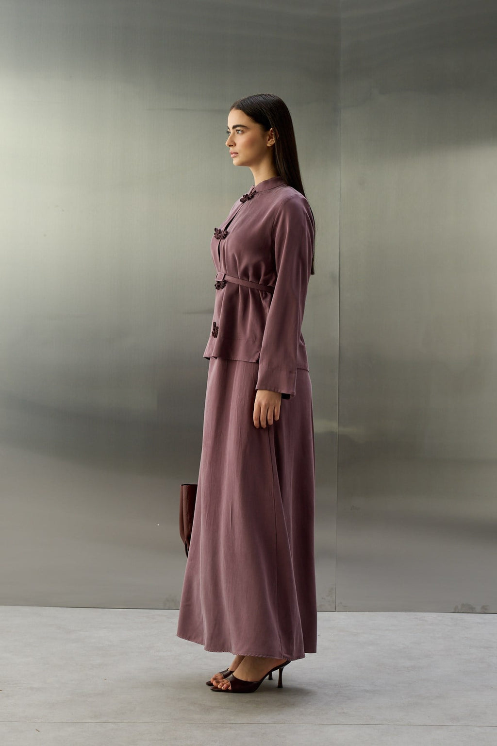 Zijaanzicht van een model met donkerbruin haar, staand voor een achtergrond van geborsteld metaal. Ze draagt een elegante, paars-lila tweedelige set van zijdeachtige stof. Het jasje heeft een opstaande kraag en donkerpaarse oosterse kikersluitingen in de vorm van gestileerde vleugels. De taille wordt benadrukt door een bijpassende riem. Ze houdt een donkerrode tas vast. Haar outfit wordt gecompleteerd door donkerpaarse, open hakken.
