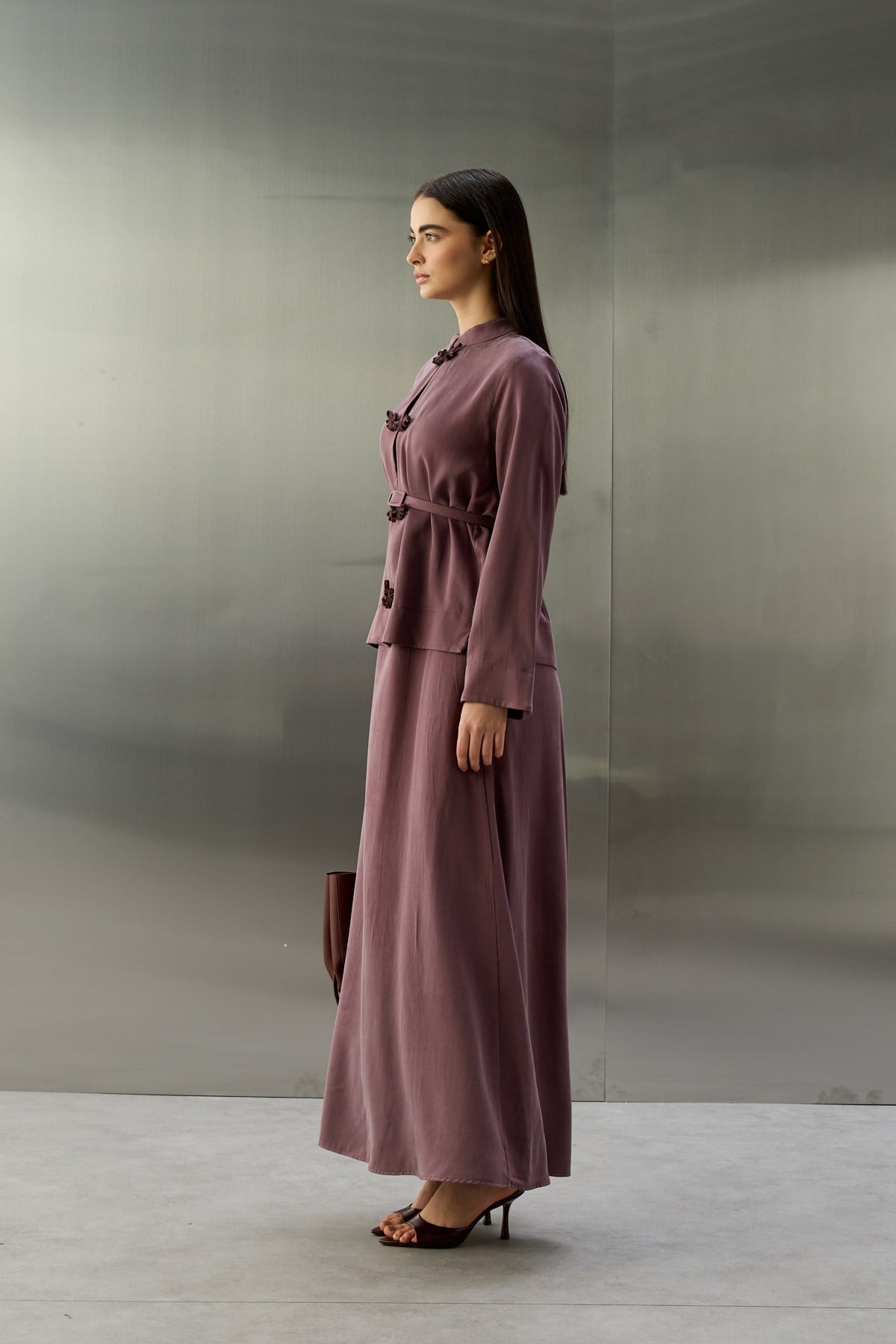 Zijaanzicht van een model met donkerbruin haar, staand voor een achtergrond van geborsteld metaal. Ze draagt een elegante, paars-lila tweedelige set van zijdeachtige stof. Het jasje heeft een opstaande kraag en donkerpaarse oosterse kikersluitingen in de vorm van gestileerde vleugels. De taille wordt benadrukt door een bijpassende riem. Ze houdt een donkerrode tas vast. Haar outfit wordt gecompleteerd door donkerpaarse, open hakken.