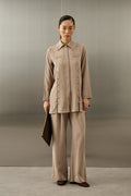 Een elegant model draagt de Serene Set in Taupe van Atelier SHÉ & CO. De set bestaat uit een zijdezachte, lange tuniek met een blinde knoopsluiting en verticale rijen ton-sur-ton sierknoopjes, gecombineerd met een bijpassende pantalon met wijde pijpen. De tuniek heeft een klassieke puntkraag en de look wordt gecompleteerd met een donkerbruine clutch en bijpassende platte schoenen.
