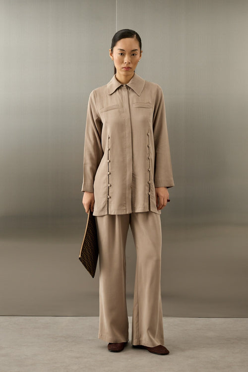 Een elegant model draagt de Serene Set in Taupe van Atelier SHÉ & CO. De set bestaat uit een zijdezachte, lange tuniek met een blinde knoopsluiting en verticale rijen ton-sur-ton sierknoopjes, gecombineerd met een bijpassende pantalon met wijde pijpen. De tuniek heeft een klassieke puntkraag en de look wordt gecompleteerd met een donkerbruine clutch en bijpassende platte schoenen.