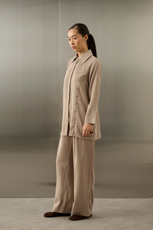 Een elegant model draagt de Serene Set in Taupe van Atelier SHÉ & CO. De set bestaat uit een zijdezachte, lange tuniek met een blinde knoopsluiting en verticale rijen ton-sur-ton sierknoopjes, gecombineerd met een bijpassende pantalon met wijde pijpen. De tuniek heeft een klassieke puntkraag en de look wordt gecompleteerd met een donkerbruine clutch en bijpassende platte schoenen.