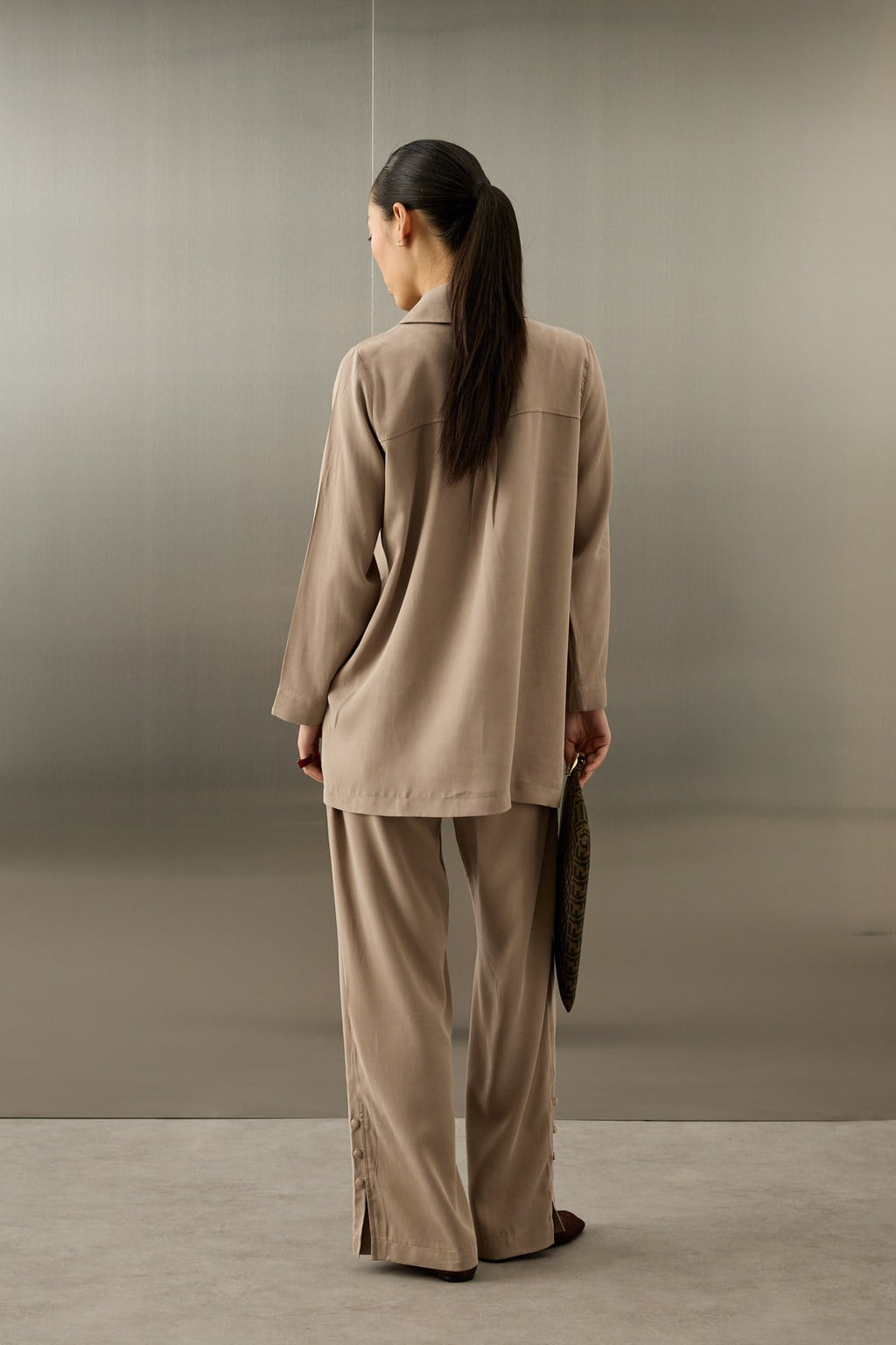 Een elegant model draagt de Serene Set in Taupe van Atelier SHÉ & CO. De set bestaat uit een zijdezachte, lange tuniek met een blinde knoopsluiting en verticale rijen ton-sur-ton sierknoopjes, gecombineerd met een bijpassende pantalon met wijde pijpen. De tuniek heeft een klassieke puntkraag en de look wordt gecompleteerd met een donkerbruine clutch en bijpassende platte schoenen.